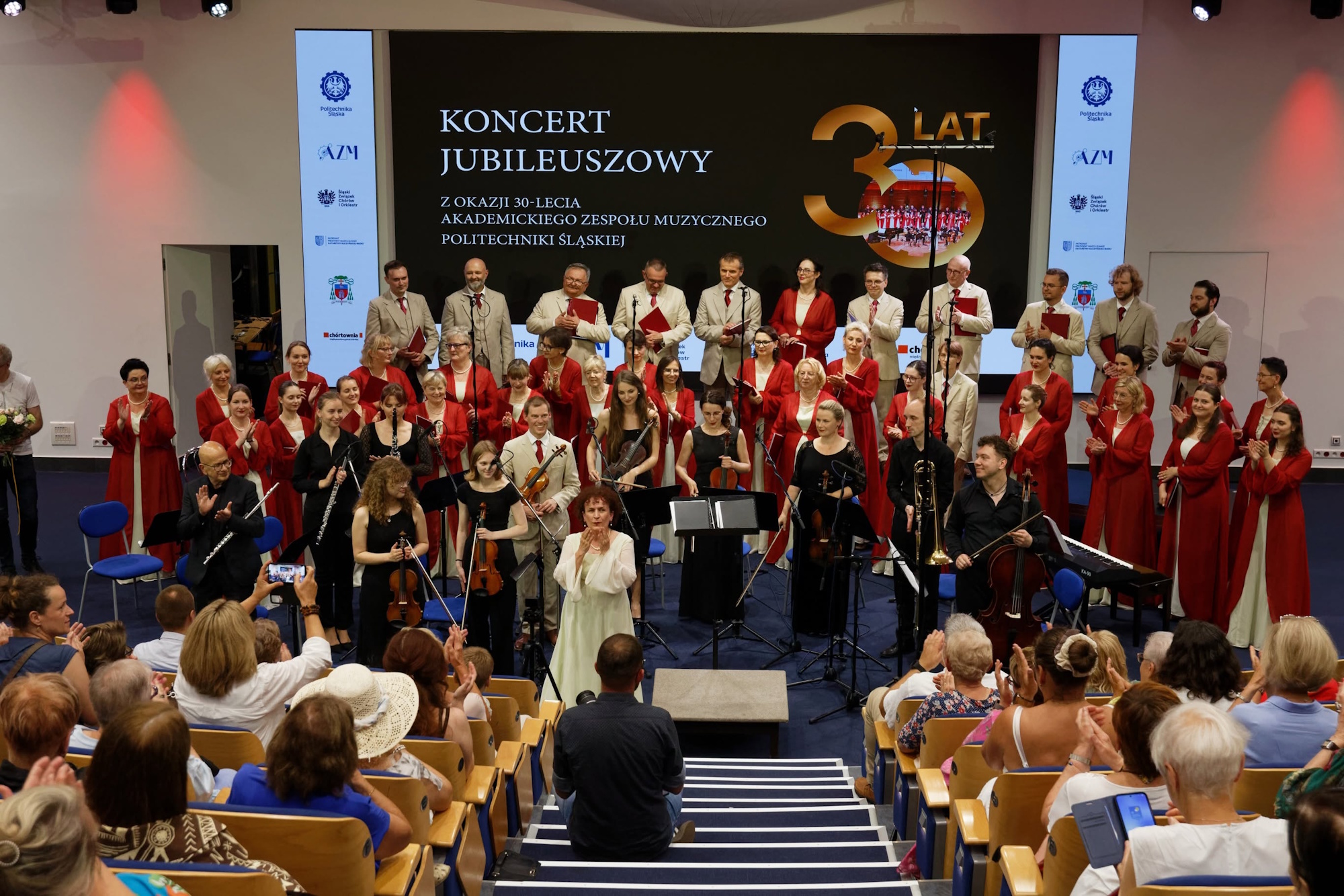 chórzyści w auli w czerwono-złotych strojach