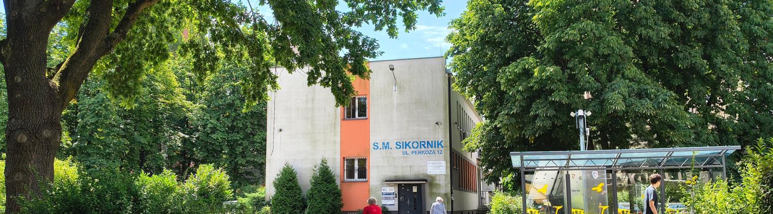 dwie kobiety z balkonikami spacerują na skwerze, obok wielkie drzewa i budynek spółdzielni